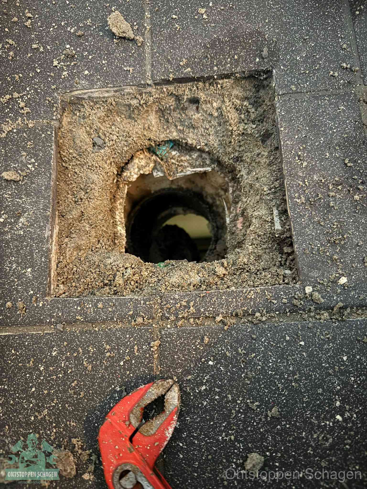 Een geopend rioolputje in een betontegelvloer met losgemaakt rooster en een zichtbare ronde afvoerbuis, het achterhalen waar de afvoerverstopping zit in Schagen