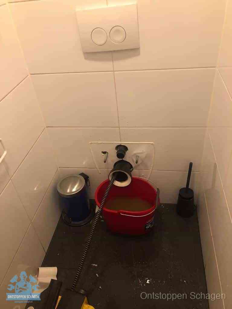 Een ontstopte wand-wc met verwijderde toiletpot waar een ontstoppingsveer in de wanduitgang verdwijnt, het achterhalen waar de verstopping zit in Schagen