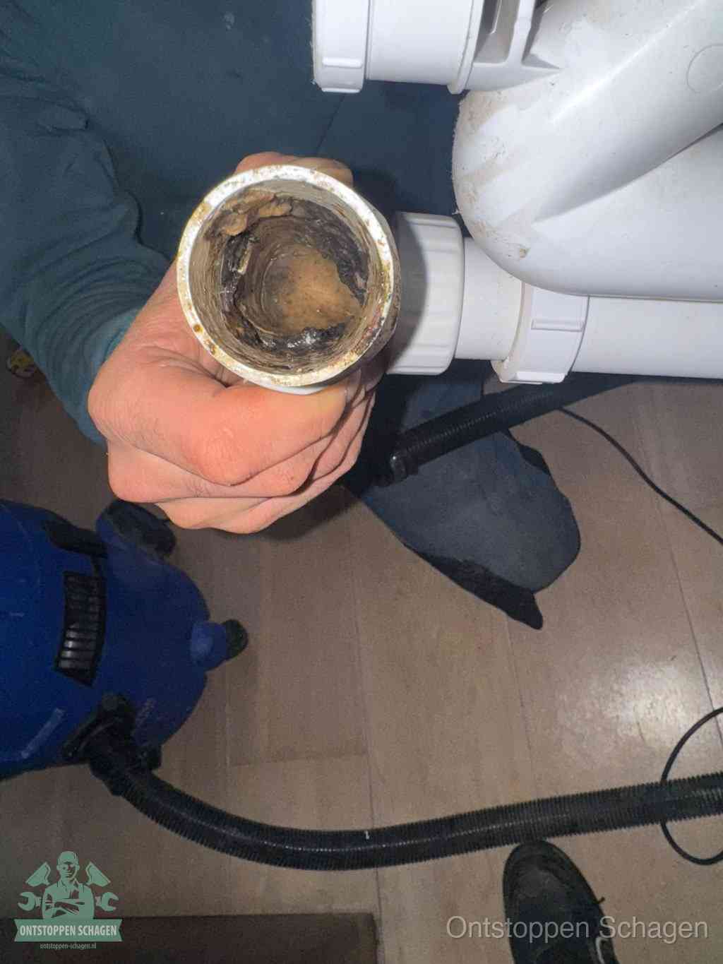 Een hand houdt een losgekoppeld PVC-afvoerbuisstuk vast dat van binnen volledig is dichtgeslibd met vet, vuil en aanslag, de meest voorkomende oorzaak van een verstopte afvoer in Schagen