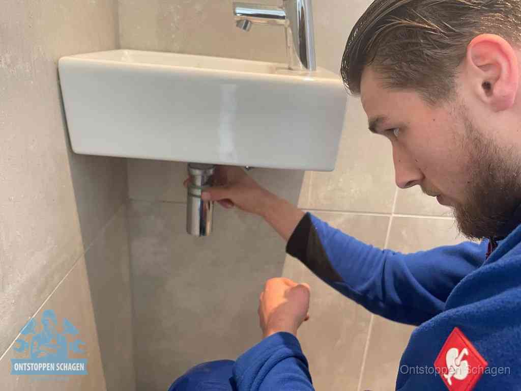 Een monteur draait met de hand de zwanenhalssifon onder een fonteinwasbak in de toiletruimte los om de bron van rioollucht in Schagen te vinden