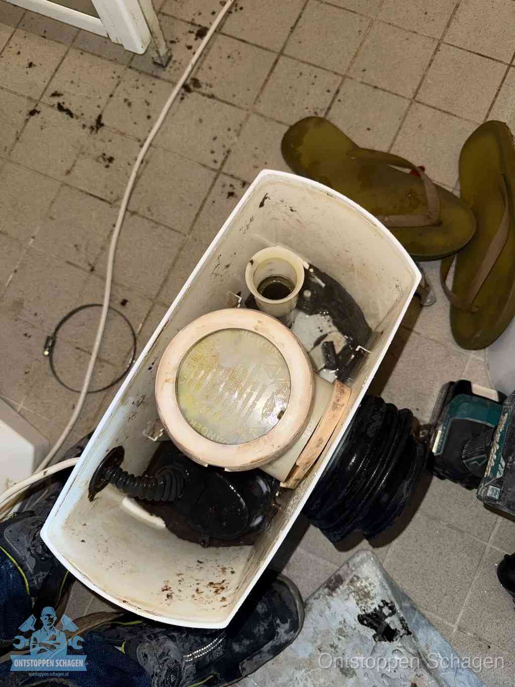 Een gedemonteerde toiletpot op de vloer met ontstoppingsveer en gereedschap ernaast, het verwijderen van een vastzittend voorwerp uit de wc in Schagen