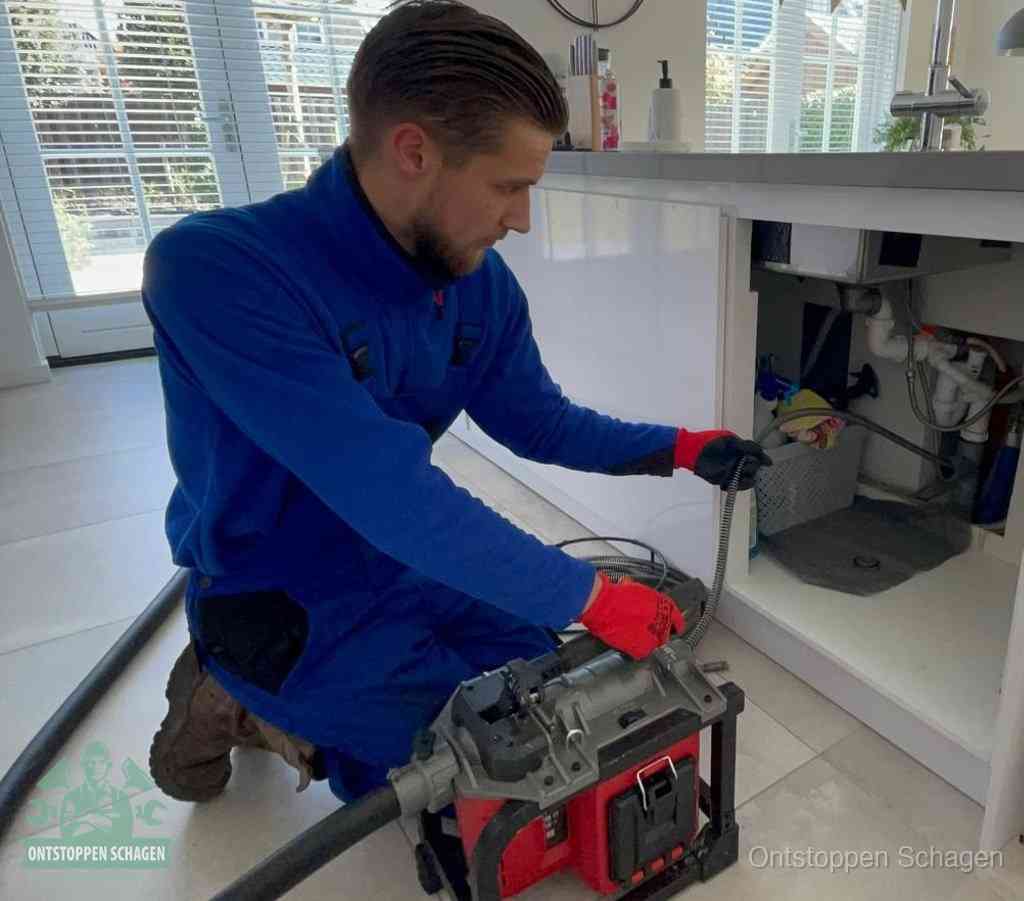 Een monteur met rode handschoenen knielt in een keuken en voert een elektrische ontstoppingsveer in tijdens het professioneel afvoer ontstoppen in Schagen